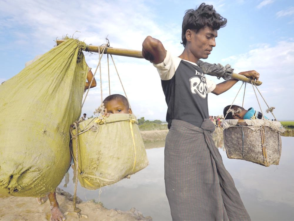 روهينجي يحمل طفليه في سلال خلال هروبه من وحشية ميانمار (رويترز)