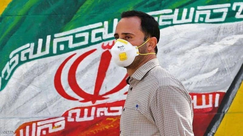 إيراني يرتدي كمامة بجوار علم لبلاده (مكة)