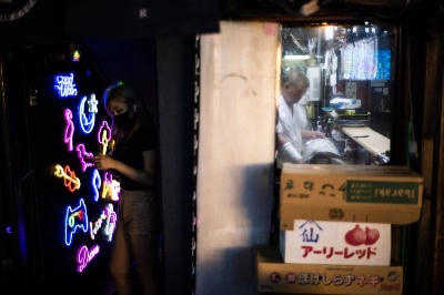 A woman stands on one side of the wall texting in front of a nightclub while, on the other side of the wall, a man works in an izakaya. A woman stands on one side of the wall texting in front of a nightclub while, on the other side of the wall, a man works in an izakaya.