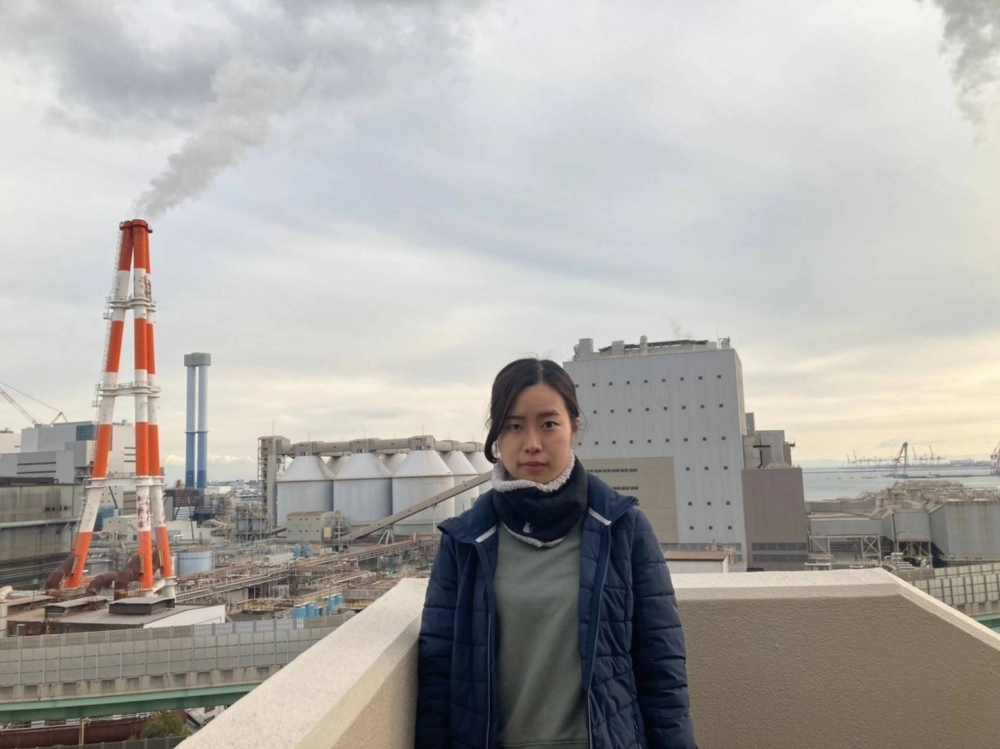 Erina Imai, a plaintiff in a climate lawsuit, poses in front of the coal-fired Kobe Power Plant in Kobe. The red and white tower behind her is private power generation equipment operated by Kobe Steel and is releasing water vapor, not smoke. Erina Imai, a plaintiff in a climate lawsuit, poses in front of the coal-fired Kobe Power Plant in Kobe. The red and white tower behind her is private power generation equipment operated by Kobe Steel and is releasing water vapor, not smoke.