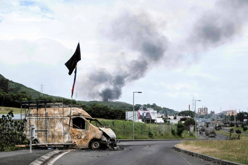 Smoke rises in the distance as a burnt vehicle blocks a road near the Montravel area of Noumea, New Caledonia, on Tuesday. Smoke rises in the distance as a burnt vehicle blocks a road near the Montravel area of Noumea, New Caledonia, on Tuesday.