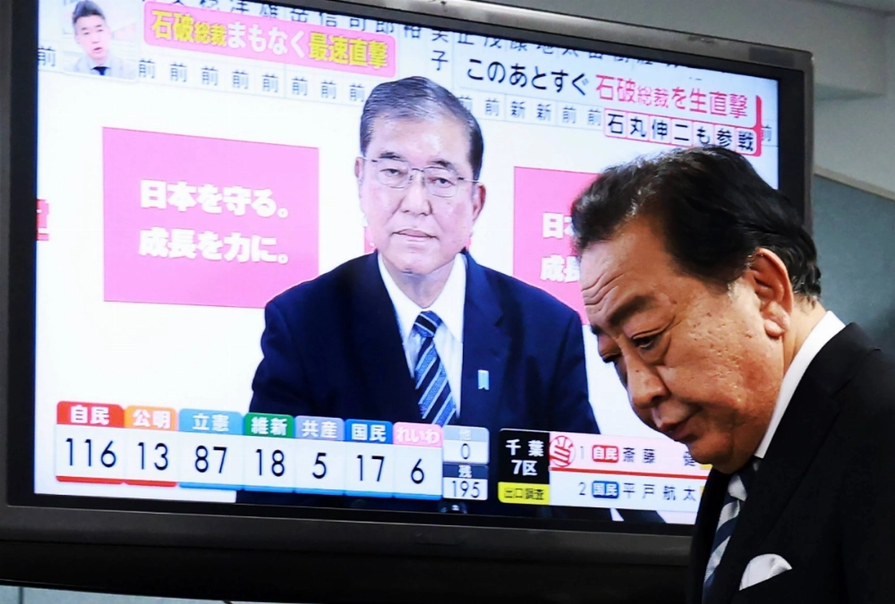 Yoshihiko Noda, head of the Constitutional Democratic Party of Japan, walks past a monitor displaying a live news broadcast showing Prime Minister Shigeru Ishiba in Tokyo on Sunday. Yoshihiko Noda, head of the Constitutional Democratic Party of Japan, walks past a monitor displaying a live news broadcast showing Prime Minister Shigeru Ishiba in Tokyo on Sunday.