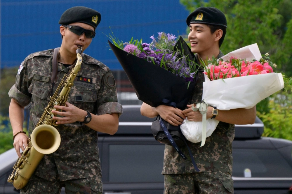 BTS member RM (left) plays saxophone while V looks on while holding flowers at an outdoor facility in Chuncheon on Tuesday.   BTS member RM (left) plays saxophone while V looks on while holding flowers at an outdoor facility in Chuncheon on Tuesday.