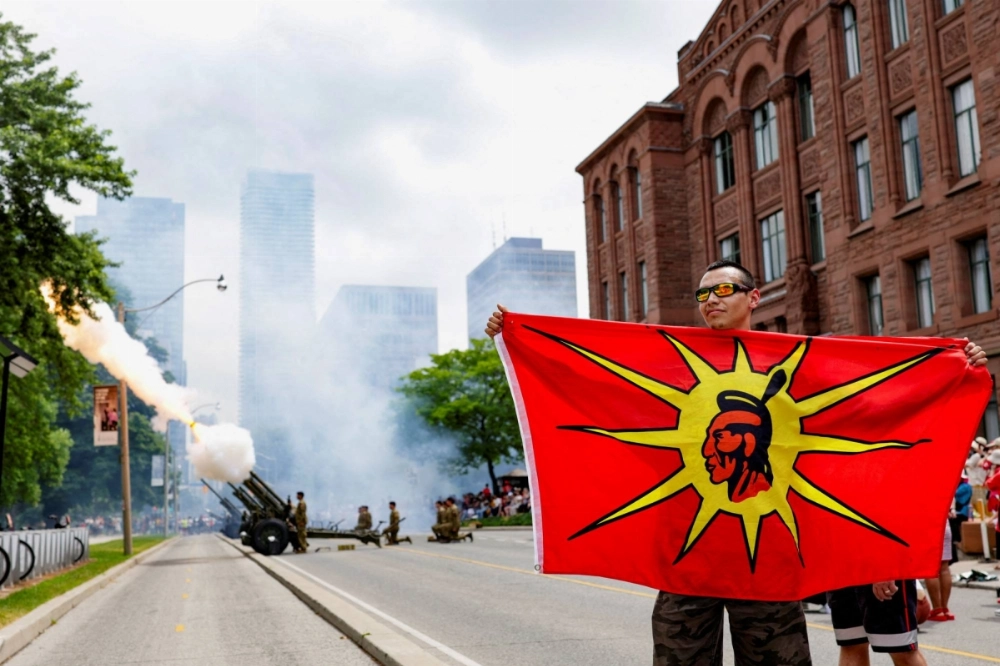 A demonstrator holds the Mohawk Warrior Flag during a protest in front of the Legislative Assembly of Ontario against the federal government’s Bill C-5, as members of the Canadian Armed Forces fire cannons in the background during a celebration marking Canada Day in Toronto on July 1. A demonstrator holds the Mohawk Warrior Flag during a protest in front of the Legislative Assembly of Ontario against the federal government’s Bill C-5, as members of the Canadian Armed Forces fire cannons in the background during a celebration marking Canada Day in Toronto on July 1.