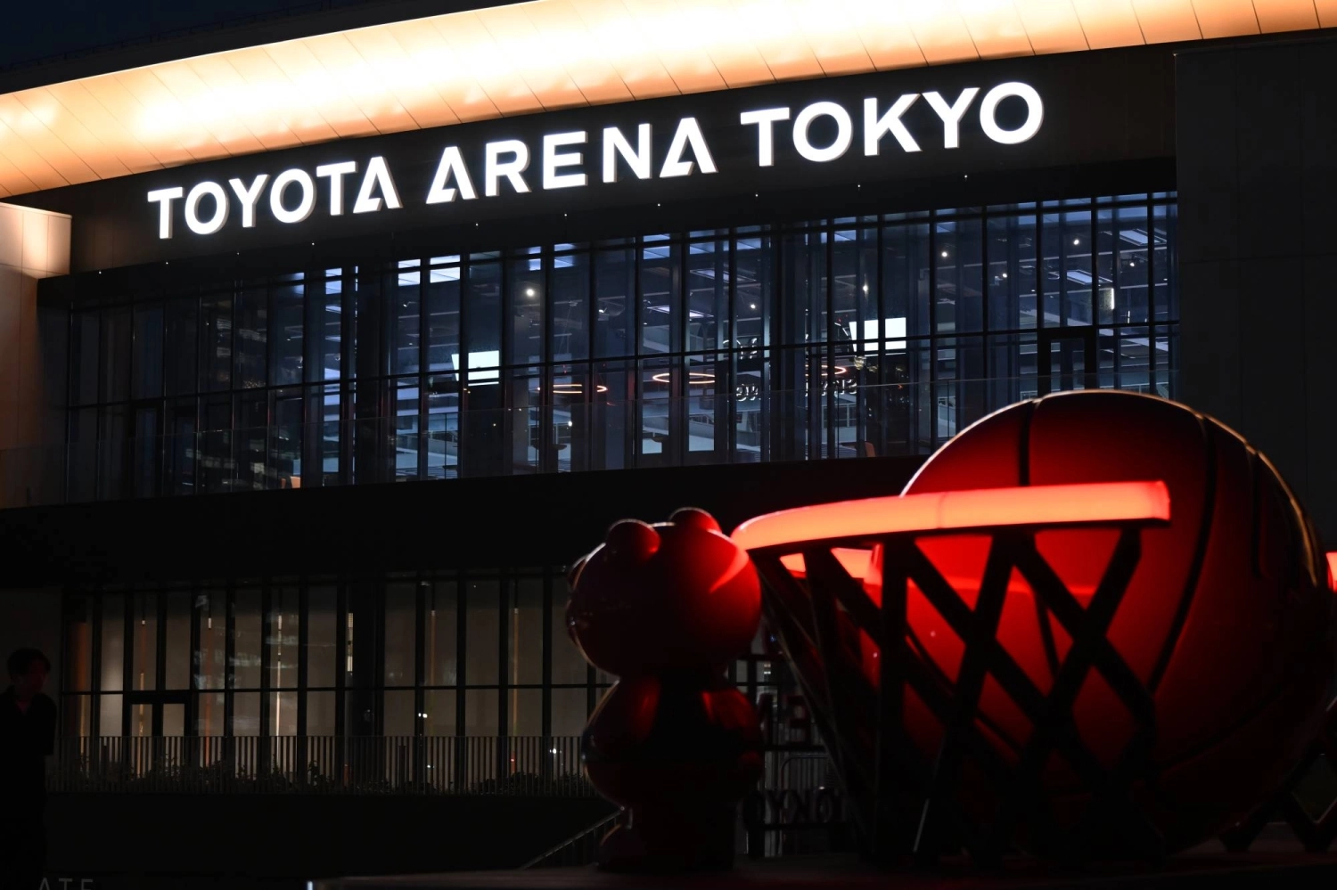Toyota Arena Tokyo lights up at night with a basketball-themed structure in the foreground. Toyota Arena Tokyo lights up at night with a basketball-themed structure in the foreground.